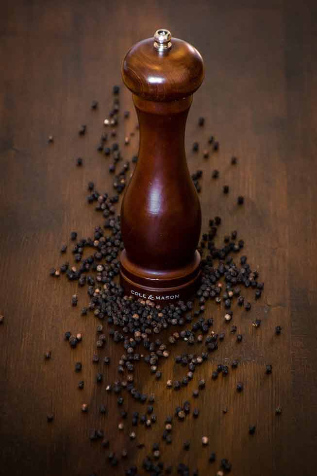 Un moulin à poivre en bois Cole & Mason, pièce maîtresse d'une table élégante, entouré de grains de poivre noir disposés pour symboliser l'Art de la table.