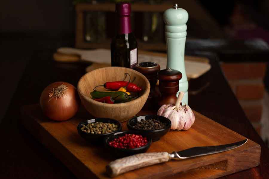 Planche en bois avec des ingrédients culinaires, comprenant des poivres de différentes couleurs dans des bols, des piments rouges dans un bol en bois, une gousse d'ail, un oignon, un moulin à poivre vert clair, une bouteille d'huile et un couteau.